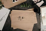 Brown rectangular pieces of paper sit askance in a pile in the center with the top one reading the word, “no” in black inked calligraphy. Surrounding the pieces of paper are various art and office materials sitting atop a gray marbled countertop.