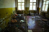 An abandoned room with peeling yellow and white paint on the walls contains several small metal bed frames, many with missing or damaged mattresses. Torn sheets, stuffed dolls, and scattered papers lie on the beds and dusty wooden floor. Light filters through large, grimy windows, casting a dim glow over the scene. The room feels still and long-forgotten, with signs of past use by children.