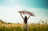 Woman standing in a field in a long dress holding a scarf above her head in the breeze