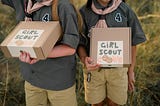 Two girls holding boxes with the words “Girl Scout Cookies” handwritten on them