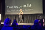 The author giving a talk on stage at an engineering leadership conference in 2023. The title slide behind her says, “Think like a journalist”