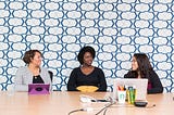 Three women sit at a long table in a work envioronment, making eye contact as if engaged in a compelling conversation.