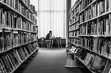 A man sitting at a table at the end of a row of books in a library.