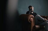 Dark photo of a man sitting on a leather couch with his head in his hands.