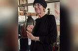 tall older white woman wearing a black top and a black knit hat. she’s holding a pigeon. she has a half smile.