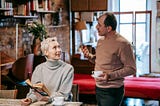 Blonde short-haired woman holding a book and talking to a man beside her, who holds a cup of coffee.