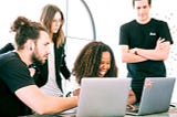Two people sitting in front of two laptops while two other people are standing behind them.