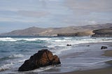 A beach on the island pf Fuerteventura, Canary Islands, Sp. A boulder is visible in the foreground, scatterered large rocks in the near distance, beach cliffs in the middle distanced and beyond those, indistinct hills.