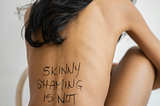 A slender woman, knees to her chest, with writing on her back reading: “skinny shaming is not okay.”
