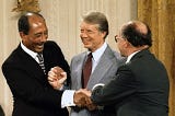 Color photograph of the three men — Egyptian President Anwar Sadat (left), U.S. President Jimmy Carter (center), and Israeli Prime Minister Menachem Begin (right) — in a standing position, shaking hands and smiling.