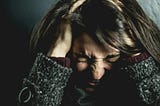 Headshot of a woman in a dark green sweater holding her head while scrunching her forehead, eyes, and nose as if in anger or frustration.