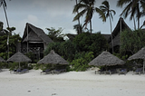 White sandy beach with Zanzibar-style thatched beach umbrellas and towering A-Frame thatched lodge fringed with palm trees