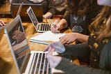 Two people sitting side by side with laptops open, conversing and writing in a notebook.