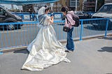 A woman standing with her back to us wearing a wedding dress and her assistant standing next to her near a railing by a busy road.