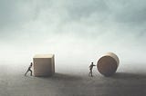 One man struggling to push a square boulder and one man pushing a round boulder with ease.