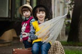 Two dark-haired boys with hats sit outside in the rain. They have a suitcase and an umbrella.