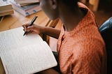 A Black woman writes with a pen in a notebook at a desk