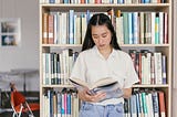 A girl is holding a book while standing in a library.