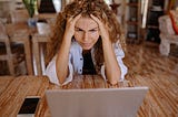 A woman with curly hair holds her head in frustration while looking at a laptop.