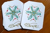 Two white cards laying on brown floorboards, with their corners cut off; the left one sits atop a small stack. The paper shows a “snowflake” design, made of green fir sprigs, red hearts, and gold embellishments. Underneath is “Warmth.” written in gold script.