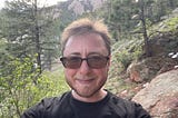 The author is standing in front of mountains, some trees, and rocks. He’s smiling. He has short brown hair and a brown beard. He’s wearing sunglasses.