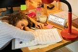 A woman asleep with her head on her desk and her elbow on an open notebook.