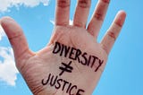 photograph of a hand with fingers outstretched and palm facing the camera. On the palm is hand-printed in black ink, “DIVERSITY ≠ JUSTICE”. In the background is a briliant blue sky with a few clouds on the left.