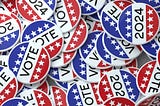 Several American VOTE buttons are scattered and piled on top of each other, each sporting white, blue, and red with white stars.