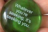 Picture is of a hand holding a green see through stone with the words “Whatever you’re seeking, it’s seeking you” written in white text.