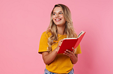 A smiling blond woman writing in a red notebook on a pink backdrop