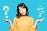 A young woman in a yellow top against a plain blue background. She is looking directly at the viewer and holding up her hands as if weighing up to options. Above each hand is a white question mark, symbolising her choices.