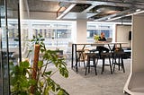 Picture of an industrial-style office with a man sitting at a high table with stools, working on his laptop. In the foreground, you can see a movable whiteboard and a monstera plant.