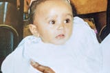 A photograph of me (Kashala, aged around 6 months or so) as a baby, dressed in white, waiting to get christened.