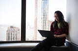 A woman working on her laptop while sitting on a window sill that overlooks a cityscape.