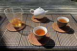 Gong Fu Cha tea set up on a black and tan criss crossed patterned placemat with a porcelain teapot, glass fairness pitcher, two black tea cups with 4 tan brown woven coasters underneath each tea ware component.