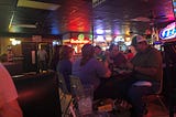 A group of six people are seated at a table in a sports bar full of neon advertisements for cheap beer (Budweiser, Miller Lite, etc.) Two are wearing matching ball caps.