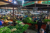 A view of a covered market hall in Kutaisi, Georgia. There are displays of fresh veggies along with vendors and shoppers.