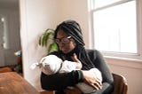 A black woman with braids and glasses holding a baby and gazing at it warmly