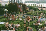 Folks picnic, talk, lounge on a large lawn with vegetable rows, flowers and a multicolor glass greenhouse in the background.