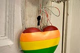 Small heart painted in the pride rainbow hanging from an old fashioned glass doorknob against a white door. Visible just above and behind the heart is a keyhole.