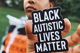 A Black child outdoors holding a sticker that reads “Black Autistic Lives Matter”