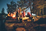 group of people sit around a campfire talking in a forest at dusk