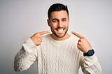 Young handsome man wearing casual sweater standing over isolated white background smiling cheerful showing and pointing with fingers teeth and mouth. Dental health concept.