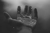 A black and white photograph of a raindrop landing in someone’s left hand.