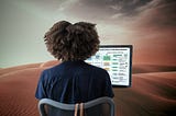 Person with big afro hair is looking at a design strategy diagram on a computer screen. In the background is a dramatic desert of red sand dunes.