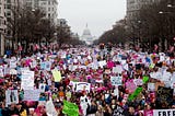A large crowd of people gathered in a peaceful demonstration in an urban area with the U.S. Capitol building visible in the background. Many participants are holding colorful signs advocating for various causes, including women’s rights, equality, and justice. The atmosphere is vibrant, with a focus on activism and solidarity. American flags and bare trees line the street, emphasizing the setting as a winter march in Washington, D.C.