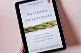 A hand holding an iPad Air tablet tilted slightly to the left, with an image of the book cover of Braiding Sweetgrass by Robin Wall Kimmerer