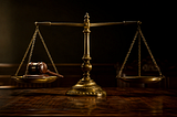 An antique brass balance scale sits perfectly level on a dark wooden judge’s bench, with a wooden gavel resting on one pan and the opposite pan empty, lit in dramatic gold and shadow.