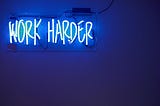 Blue neon sign on a dark wall displaying the motivational phrase “WORK HARDER” in capital letters.