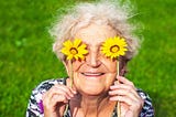 A cheerful grandmother playfully holds yellow daisies up to her eyes.